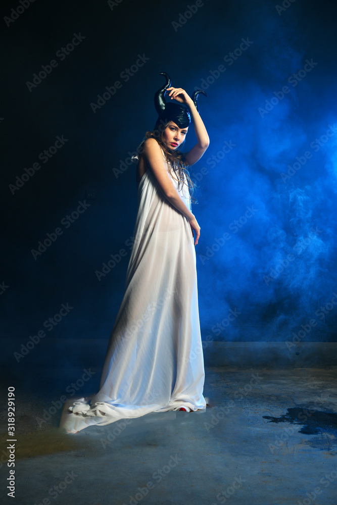 beautiful girl in a black helmet with horns and in white clothes on a blue background in smoke beautiful girl in a black helmet with horns and in white clothes on a blue background in smoke