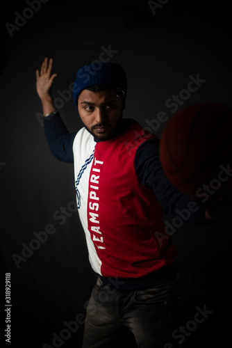 Fashion portrait of an young Indian Bengali brunette man in casual tee shirt and head band playing with basket ball in black copy space background. Indian lifestyle and fashion photography. © abir