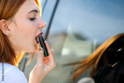 Closeup of a young redhead woman driver correcting her makeup with dark red lipstick looking in car reflective window.