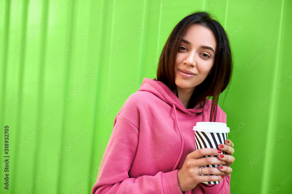 Stylish happy young woman wearing pink sweatshirt holding on the go. Portrait of smiling girl drinking coffee outdoors on the green background