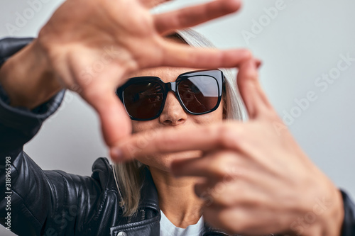 Beautiful woman in glasses, in a black leather jacket on a gray background, making a frame using hands with palms and fingers, camera perspective.