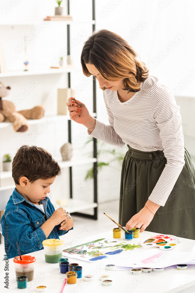 young mother standing near table while cute son holding container with paints young mother standing near table while cute son holding container with paints