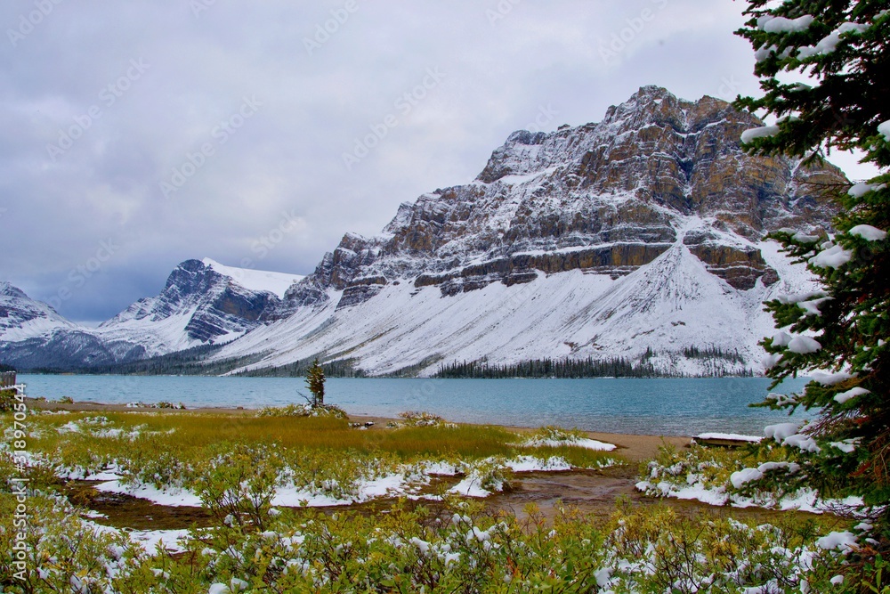 Blue water of Bow Lake, mountains covered with snow. Wooden Bridge, gray clouds, view from shore. Banff National Park, Rocky Mountains, Canada.