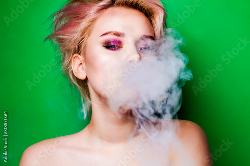 Portrait of a young smoking woman on a green background. A woman with a bright makeup makes smoke and looks into the frame.