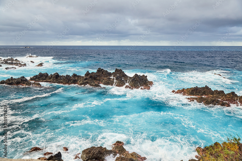 Shot of the rocky atlantic coast of Madeira island, Portugal with restless, clear, blue water and a lot of foam at the foot of the cliff and rocks