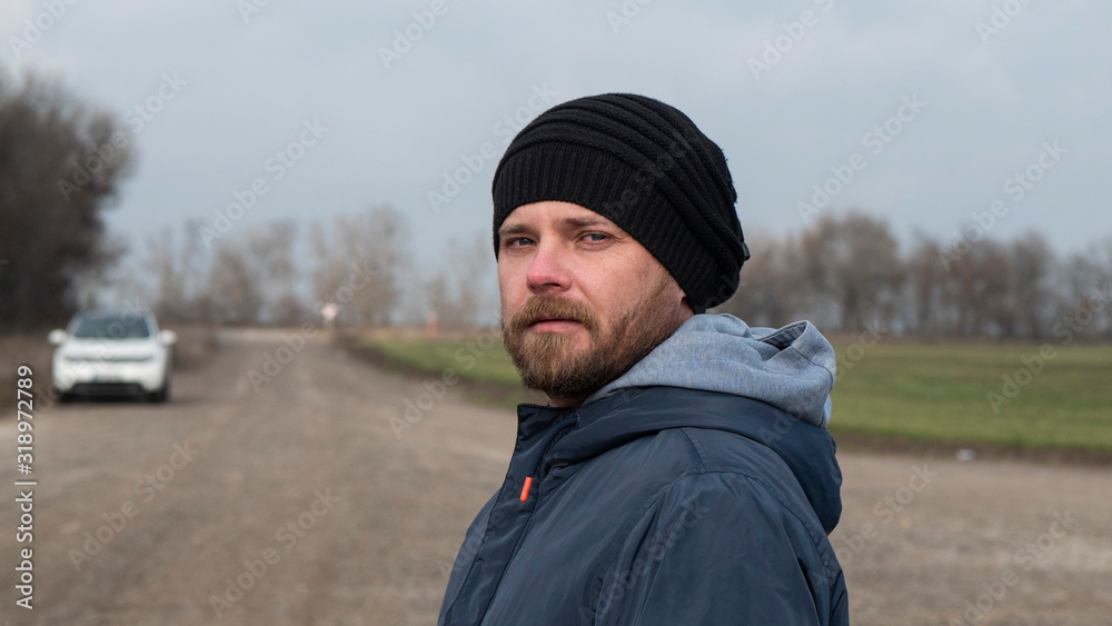 Portrait Of An Attractive Hipster Man With A Beard In A Knitted Hat. Man Against Blurry Car Background Portrait Of An Attractive Hipster Man With A Beard In A Knitted Hat. Man Against Blurry Car Background