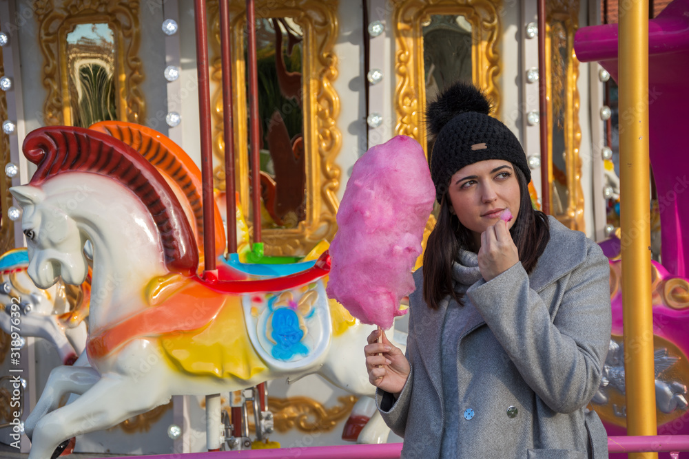 Medium shot of young brunette woman, looking to the right, with pink cotton candy, in front of a carousel, with sunset light, horizontally in Madrid, Spain