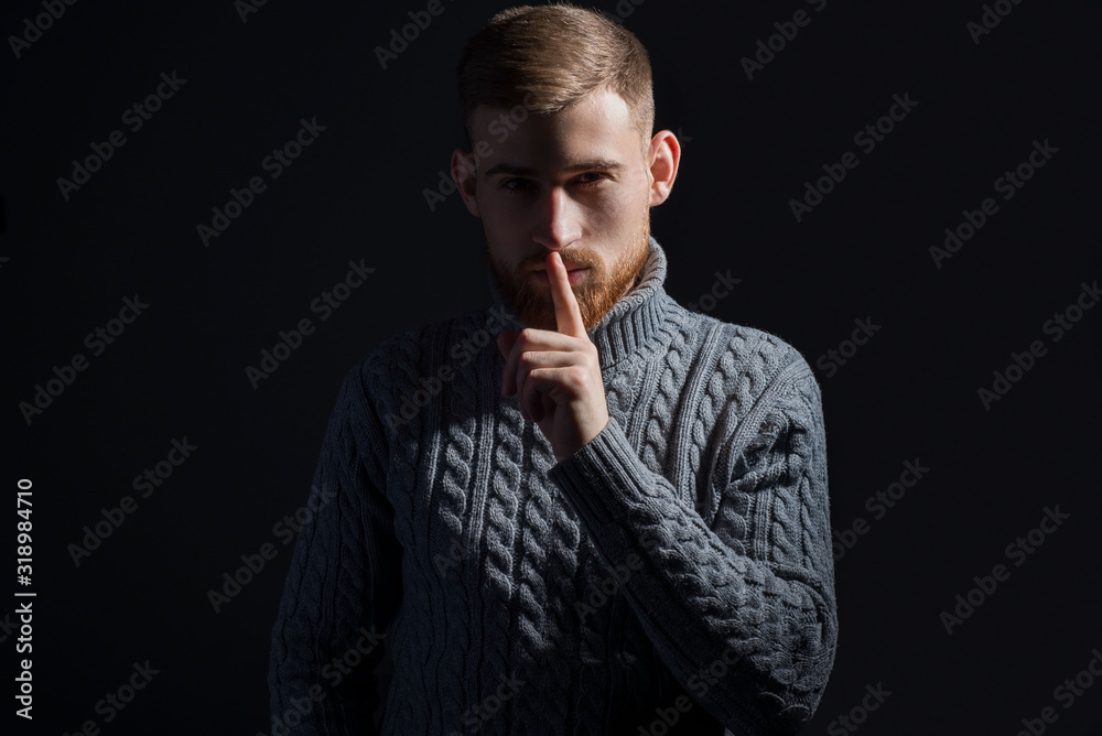 portrait of a young bearded guy of twenty-five years old. In a warm winter sweater, proudly, gray, looking at the camera and holding a finger to his mouth, with the background for a call to silence. portrait of a young bearded guy of twenty-five years old. In a warm winter sweater, proudly, gray, looking at the camera and holding a finger to his mouth, with the background for a call to silence.