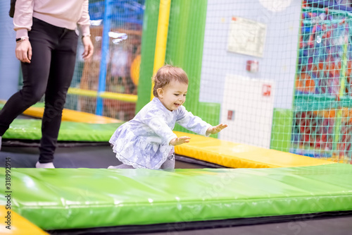little cute toddler girl in a dress with parent jumping on a trampoline and laughing. on the children's playground in the children's play center. soft focus. blurred background