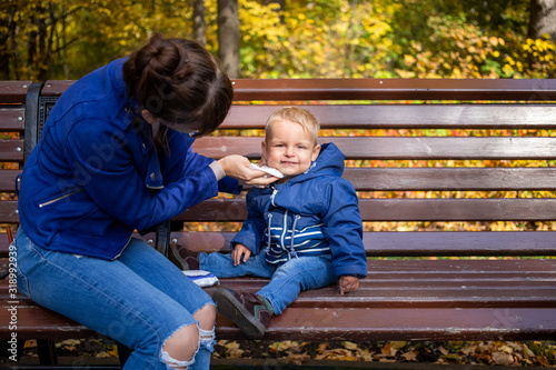 little cute baby in blue clothes sits on a bench in the park, looks at the camera and smiles, while Mom wipes his dirty face with a napkin. close-up, on background are autumn trees in blur