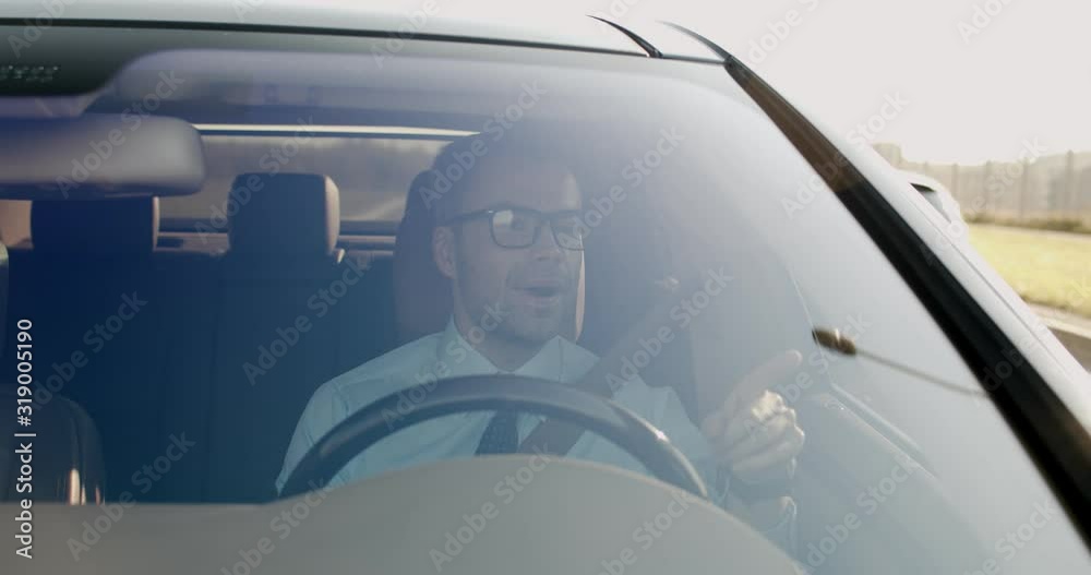 View from the outside of the car on the Caucasian good looking man in glasses driving his car on the way to work in the morning and singing. View from the outside of the car on the Caucasian good looking man in glasses driving his car on the way to work in the morning and singing.