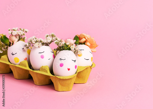 Easter creative concept. Easter decorative eggs with painted cheerful faces and flowers instead of hair in the cardboard egg tray on the pink backgraund. Copy space, close up
