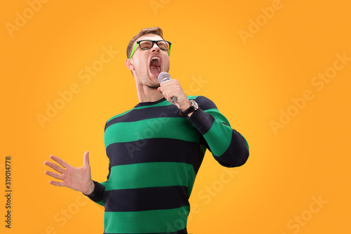 Young man in glasses singing song in microphone. Young posing isolated on yellow orange background. People lifestyle concept.