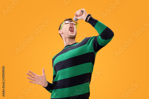 Young man in glasses singing song in microphone. Young posing isolated on yellow orange background. People lifestyle concept.