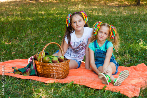 Two Girls Sit On Orange Picnic Blanket With Picnic Basket In The Park