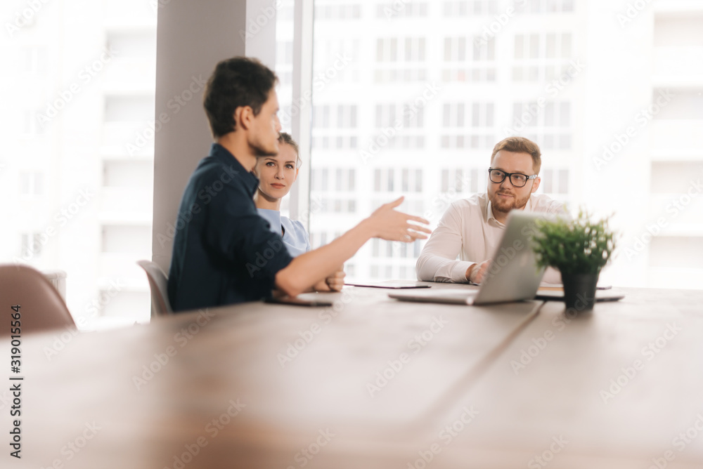 Young business man tells his partners about the project in modern office meeting against large panoramic window. Young man and woman listen to the speaker. Concept of office life. Young business man tells his partners about the project in modern office meeting against large panoramic window. Young man and woman listen to the speaker. Concept of office life.