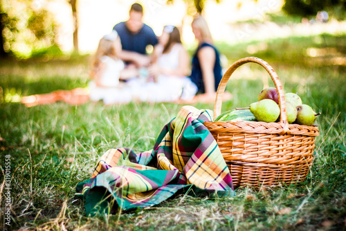 Picnic Basket With Blanket and Pears On The Grass. On Front Of The Family In Bokeh