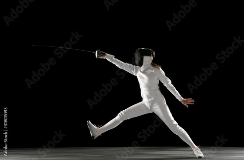 Targets. Teen girl in fencing costume with sword in hand isolated on black background. Young female model practicing and training in motion, action. Copyspace. Sport, youth, healthy lifestyle. © master1305
