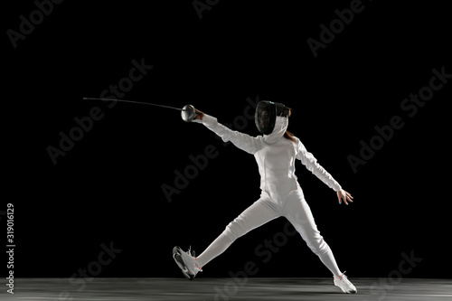 Targets. Teen girl in fencing costume with sword in hand isolated on black background. Young female model practicing and training in motion, action. Copyspace. Sport, youth, healthy lifestyle.