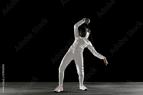 Catching moment. Teen girl in fencing costume with sword in hand isolated on black background. Young female model practicing and training in motion, action. Copyspace. Sport, youth, healthy lifestyle.