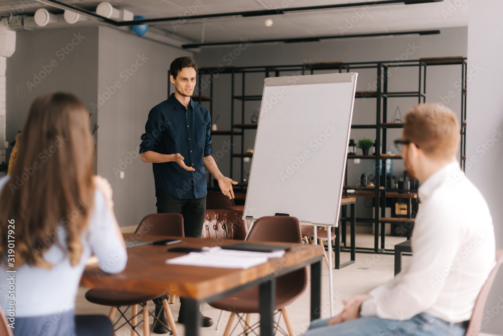 Confident young speaker coach gives corporate presentation on whiteboard in modern office meeting. Team leader teaching sales at group meeting. Male business trainer presenter speaking at training.