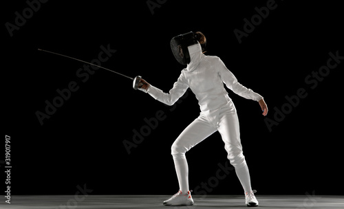 Invincible. Teen girl in fencing costume with sword in hand isolated on black background. Young female model practicing and training in motion, action. Copyspace. Sport, youth, healthy lifestyle.