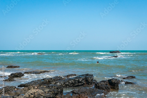 The shore of Arabian sea coast in India  Goa. Large boulders on the shore. Surf Waves breaking on the stones. Horizon line of the ocean. A young man  in waves. Dangerous swimming. 