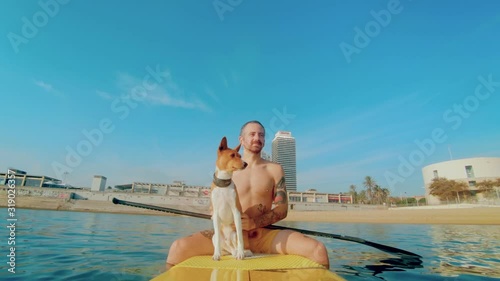 Handsome surfer man with body tattoos sit on top of surf board or sup paddle on sunny summer day. Cute funny dog in water. Concept colorful holiday lifestyle. Vacation with pet, brown dog