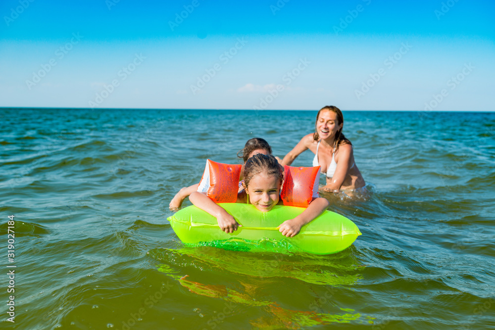 Happy mother and two small positive daughters bathe and swim in the sea with an air mattress on a sunny summer day. Vacation concept with kids. Copyspace