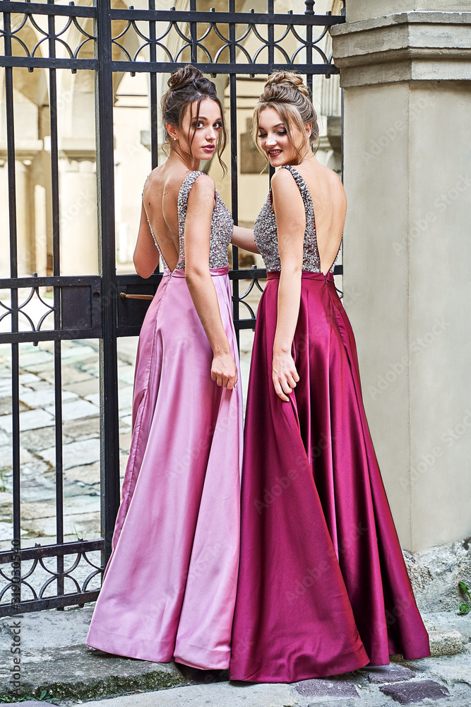 Fototapeta premium Two beautiful bridesmaids girls blonde and brunette ladies wearing elegant full length purple violet lilac lavender satin folded bridesmaid dress with silver sequined camisole top with sequins