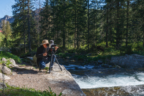 Travel photographer man in beige hat with professional film camera on tripod shooting wildlife of mountains forest river landscape Hiker tourist professional photography shooting, filming backstage