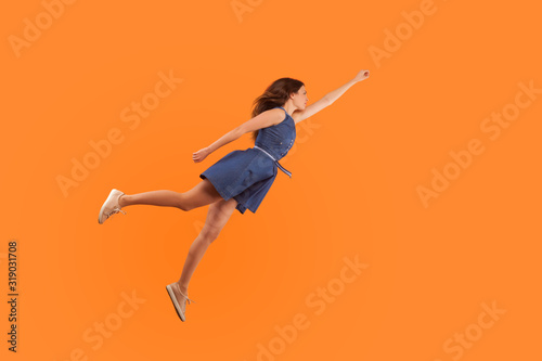 Superhero. Full length of confident motivated brunette woman in denim dress flying in air like superman and feeling superpower, striving up for success, reaching goal. studio shot orange background