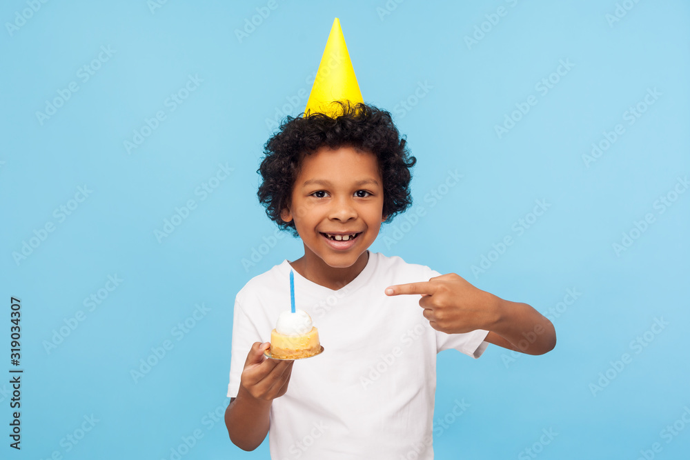 Φωτογραφία Little adorable happy boy with funny party cone on head pointing at cake with candle and looking at camera with toothy smile, expressing pure child happiness