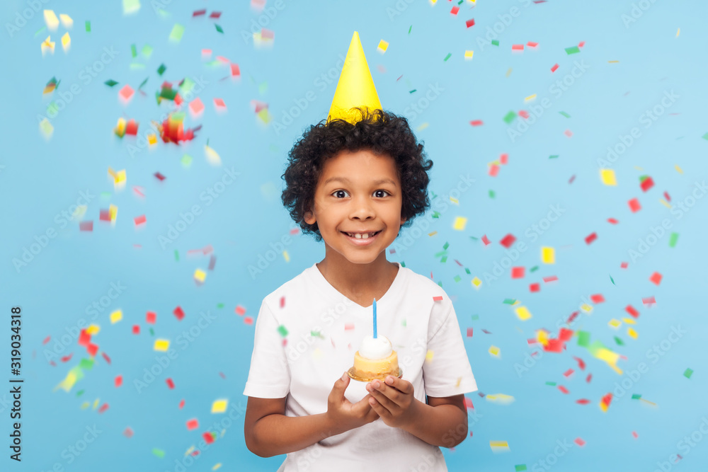Happy cheerful cute little boy with funny party cone on head holding cupcake and smiling while confetti falling around, his look expressing pure joy and happiness. indoor studio shot blue background - obrazy, fototapety, plakaty 