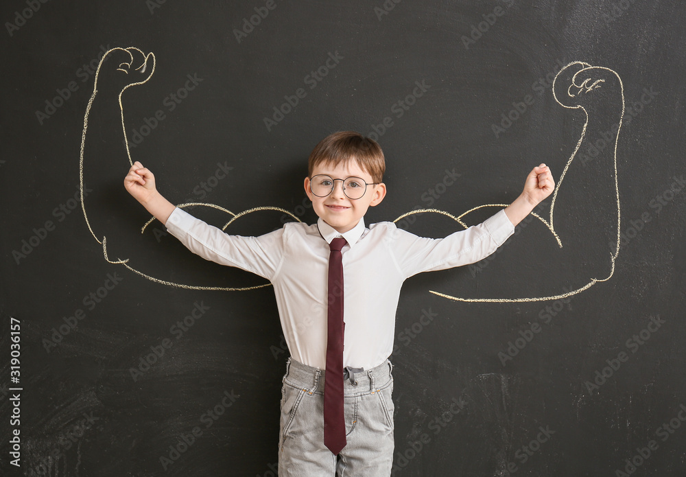 Little schoolboy near blackboard with drawn muscular arms in classroom