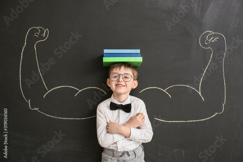 Little schoolboy near blackboard with drawn muscular arms in classroom