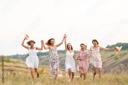 The company of cheerful female friends have a great time together on a picnic in a picturesque place overlooking the green hills. Girls in white dresses dancing in the field © Kate