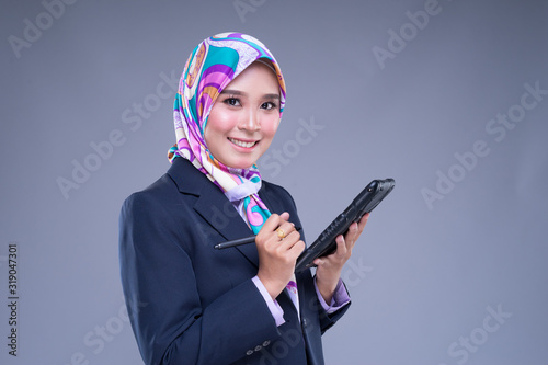 Half length portrait of an attractive Muslim woman wearing business attire and hijab poses holding a tablet isolated on grey background. For image cut-out for technology, business or finance. © HEMINXYLAN