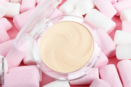 Beige face powder in a transparent package on a pastel pink background with sweets. Nude basic face makeup concealer on white marshmallows. Women's cosmetics attribute photo