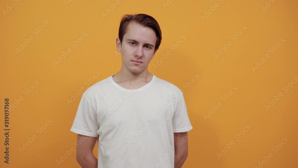 Young man in white t-shirt on yellow background has his hands behind back and looks confidently at camera ahead Young man in white t-shirt on yellow background has his hands behind back and looks confidently at camera ahead