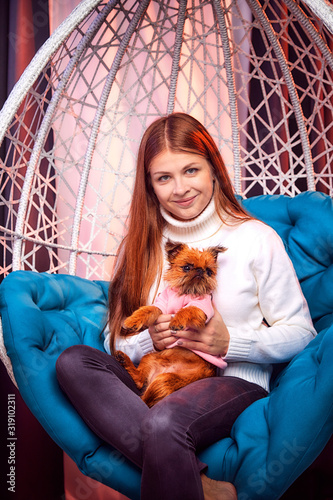 Beautiful brunette girl whth long hair in the blue shirt and small dog on a white wicker hanging chair with pillow in the room with curtains. Model during photoshoot photo