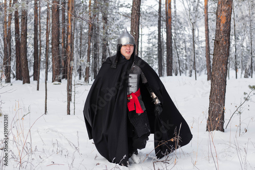 Medieval warrior in armor, helmet, black cloak with a saber in his hands in the winter in the forest.