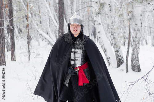 Medieval warrior in armor, helmet, black cloak with a saber in his hands in the winter in the forest.