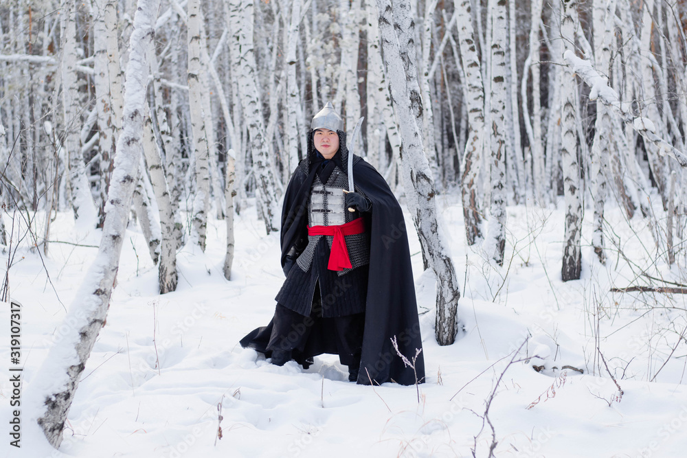 Medieval warrior in chain mail, helmet, black cloak with a saber in his hands against the background of a winter birch forest.