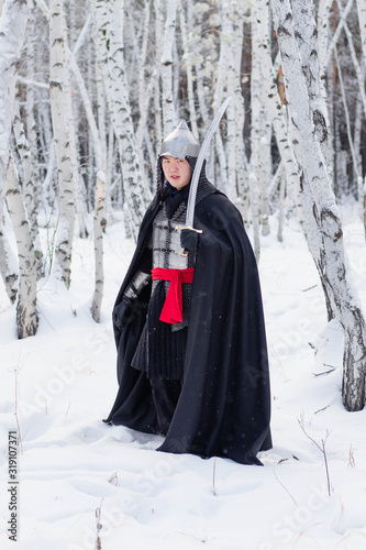 Medieval warrior in chain mail, helmet, black cloak with a saber in his hands against the background of a winter birch forest.