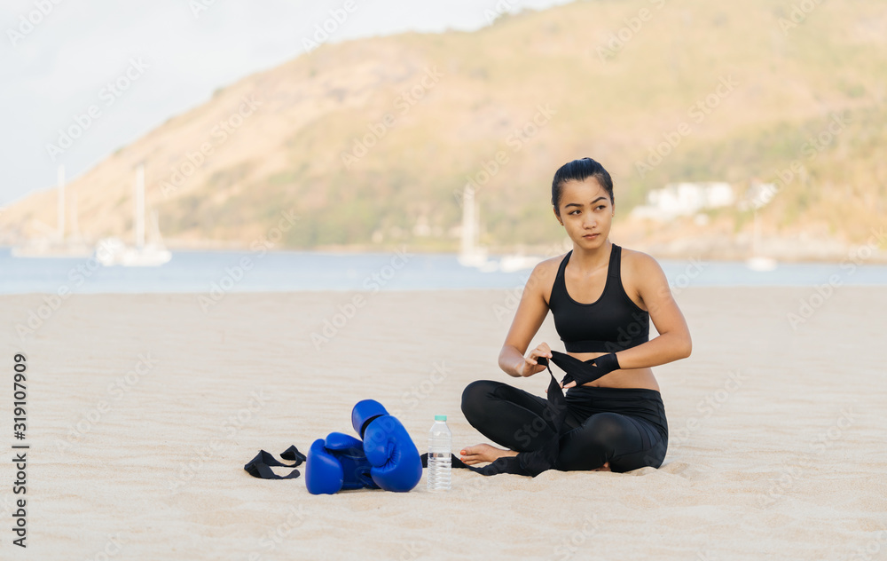 A beautiful young girl of Asian appearance in sportswear is sitting on the sand of a beach and getting ready for a boxing training. Healthy lifestyle and sport.