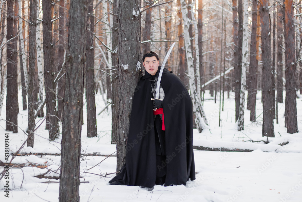 A medieval warrior in chain mail armor, a helmet and a black cloak with a saber in his hands, stands aside to a tree. Against the background of winter forest and snow. A medieval warrior in chain mail armor, a helmet and a black cloak with a saber in his hands, stands aside to a tree. Against the background of winter forest and snow.