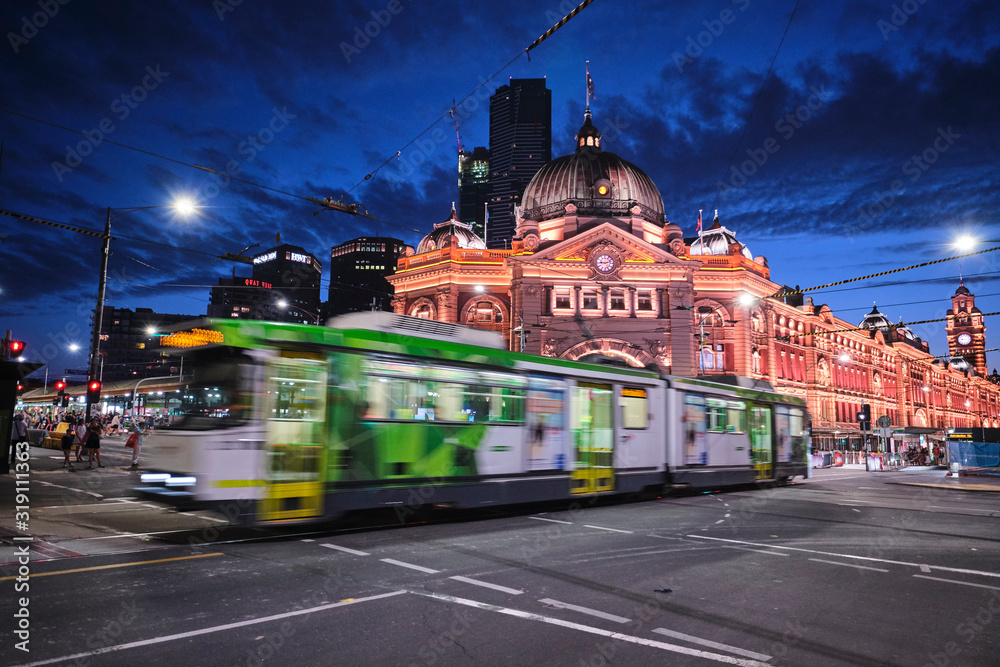 Naklejka premium Melbourne, Wiktoria / Australia - 26 stycznia 2020 r .: Stacja Flinders Street i kościół z lekkimi szlakami, szlakami samochodowymi, tramwajami, samochodami i turystami o niebieskiej godzinie