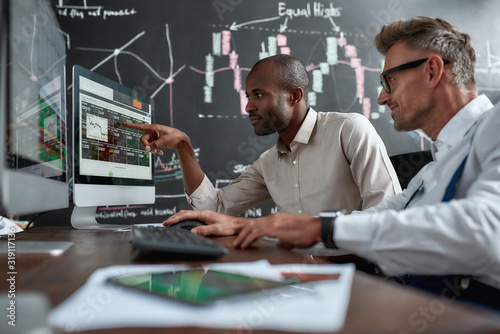 Nourishing Your Dreams. Two diverse colleagues traders discussing ideas while sitting in the office in front of multiple computer screens. Blackboard full of charts in the background.