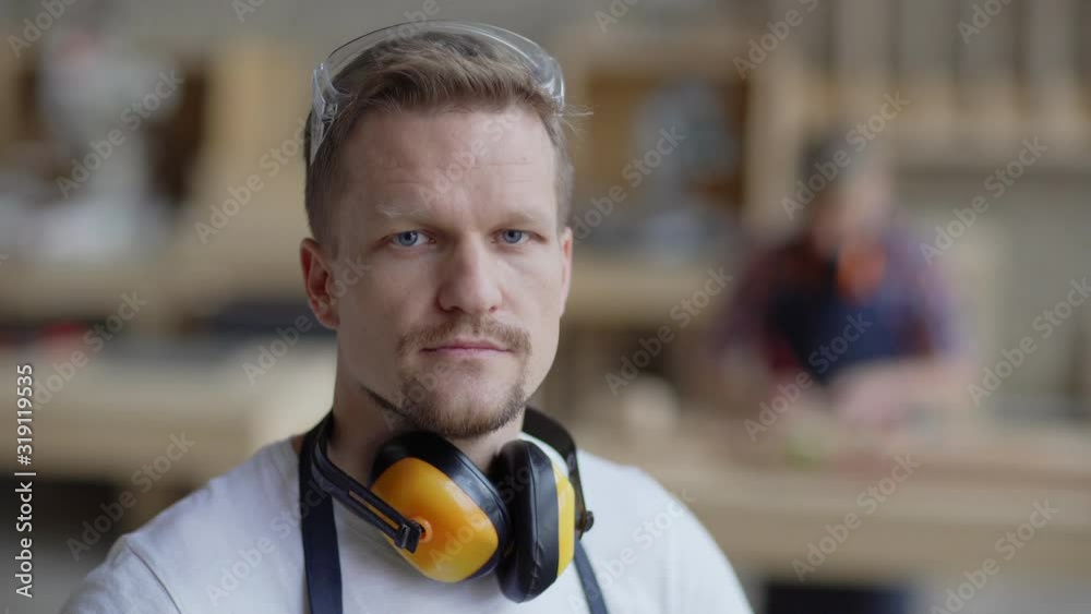 Head and shoulders video portrait of serious middle aged carpenter looking at camera in carpentry shop with protective earmuffs around his neck while smb woodworking in background, copy space to right
